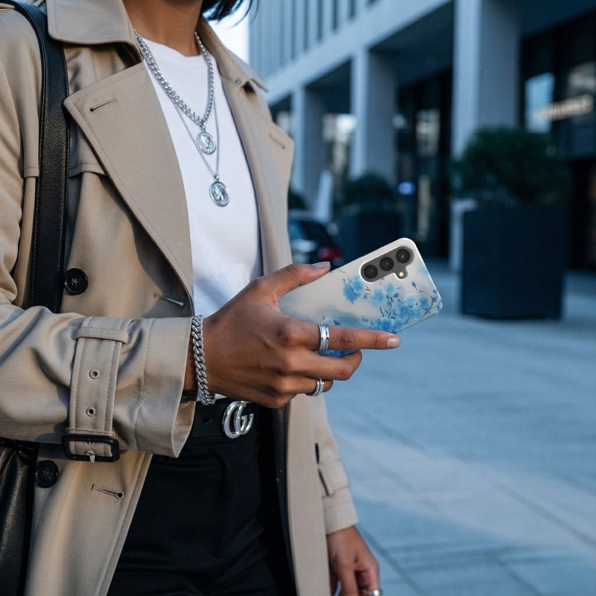 Nahaufnahme einer Person in einem beigen Trenchcoat, die ein Smartphone mit einer blau-weißen Handyhülle im Kirschblüten-Design hält, urbaner Hintergrund in einer modernen Stadt.